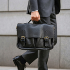 A man in a gray suit carrying a black pebbled leather briefcase with gold-tone hardware, shown from the waist down against a light gray stone wall.