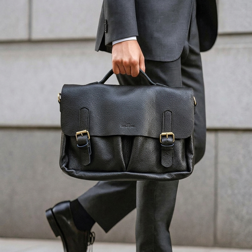 A man in a gray suit carrying a black pebbled leather briefcase with gold-tone hardware, shown from the waist down against a light gray stone wall.